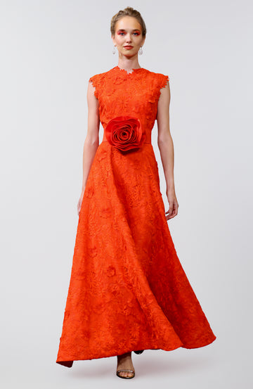 Elegant woman wearing a bold red-orange lace gown with floral embroidery, featuring cap sleeves and a statement oversized rose belt at the waist, paired with delicate earrings and open-toe heels.
